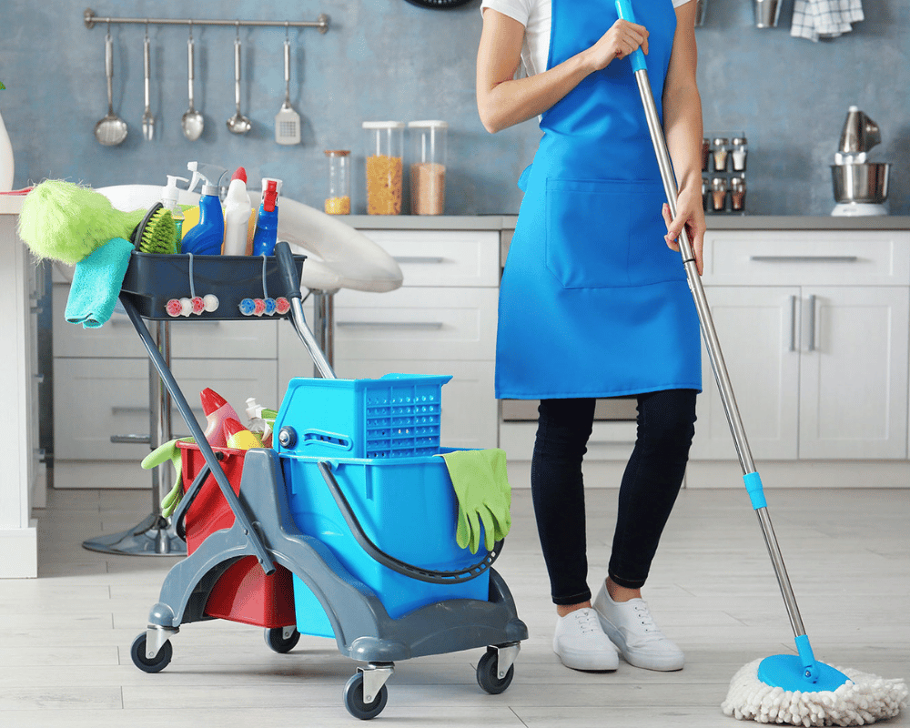 A woman in an apron holding a mop next to cleaning supplies.