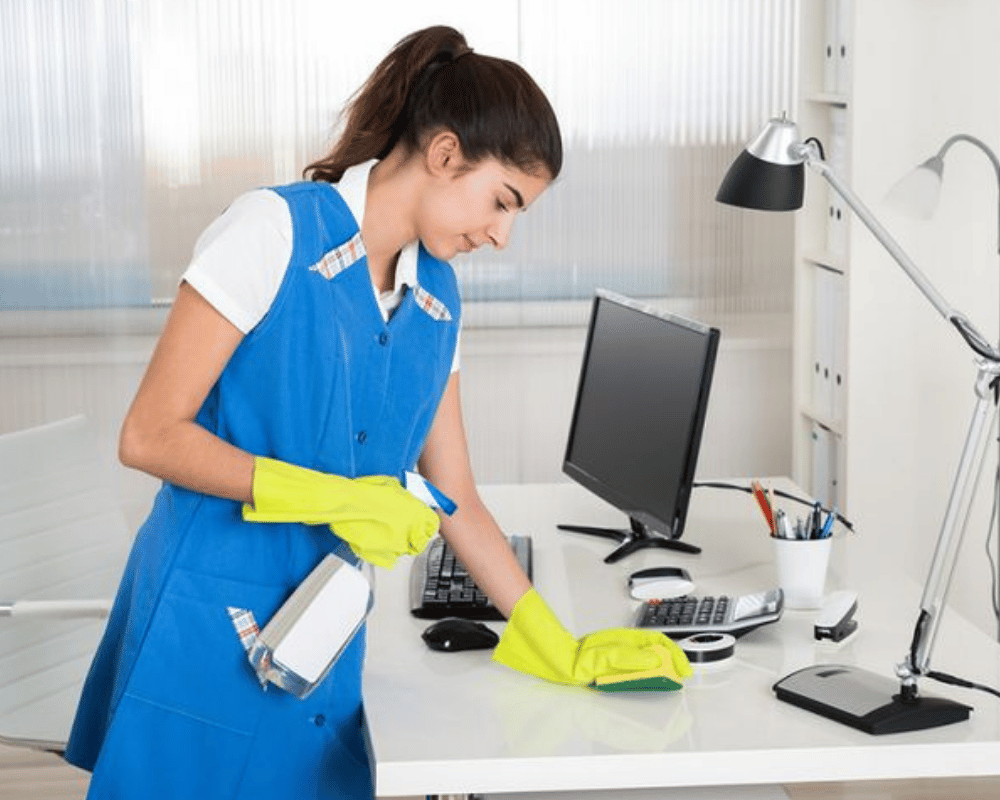 A woman in blue shirt and yellow gloves cleaning desk.